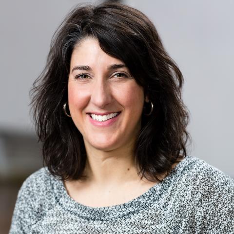 a white woman with shoulder length dark hair wearing a grey top.