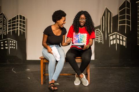 Two young women sit on a stage reading lines in front of an illustrated cityscape.