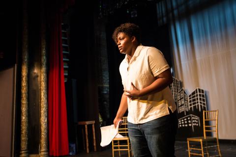 A young person delivers an impassioned speech on a stage, with their hand in a fist by their core.