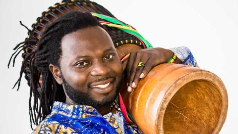 Theo is a black man with shoulder length braids. He poses with a bongo over his shoulder.