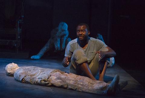 A man dressed in a t shirt, rolled up pants, and no shoes sits on a darkened stage. A prone figure is on the floor in front of him; other figures are in the background.