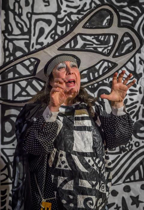 A woman in a black and white graphic print costume and hat with her hands in front of her. A bold graphic black and white wall hanging is in the background.hanging