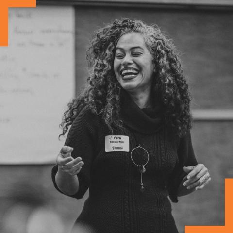 In front of large pads with text written on them, Yara smiles. Yara has thick curly hair and wears a sweater dress with her nametag on her chest.