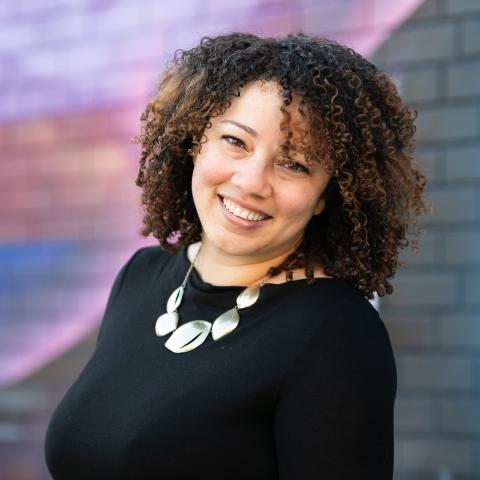 A lights-kinned woman smiles. She has shoulder length curls and wears a metallic necklace over a black blouse.