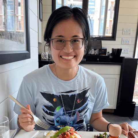 An Asian person sits a table with food in front of them. They smile and wear big eye glasses with thin frames.