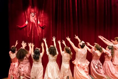 From a red curtain, a man in drag is spotlight. Before the curtain, a crowd of men and women in pink dresses hold their arms up.
