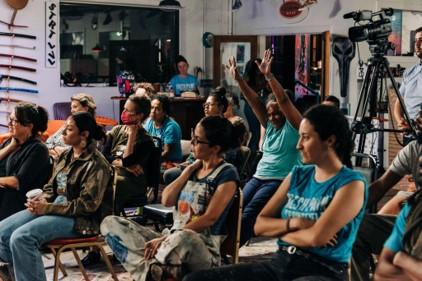 Indoors, a group of folks, some in matching turquoise t-shirts, sit and look in the same direction.