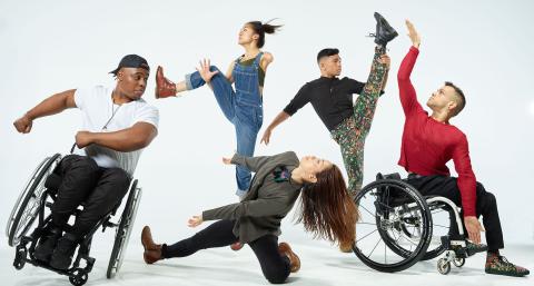 Five dancers, two of which are in wheelchairs, dance in front of a white backdrop.