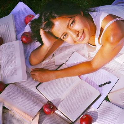 Chavi, who leans over books and red apples while laying in grass, looks up and smiles.