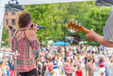 The back of a guitarist and a woman singing into a microphone on an outdoor stage in front of a crowd and summer trees.