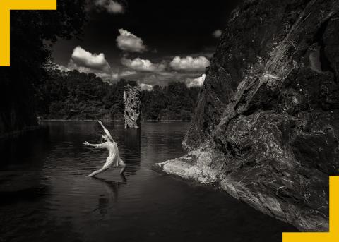 Black and white: a white woman throws her arms back and leans back next to a mountain.