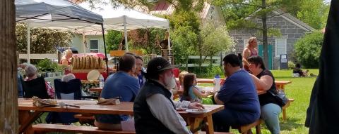 a group of people sitting at picnic tables in a green landscape. several artist display tents are are surrounding the people.