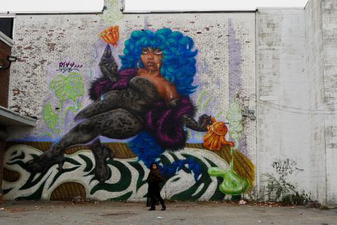 A woman of color poses in front of a mural of a woman of color, with blue hair, a purple feather boa, and a leopard catsuit.
