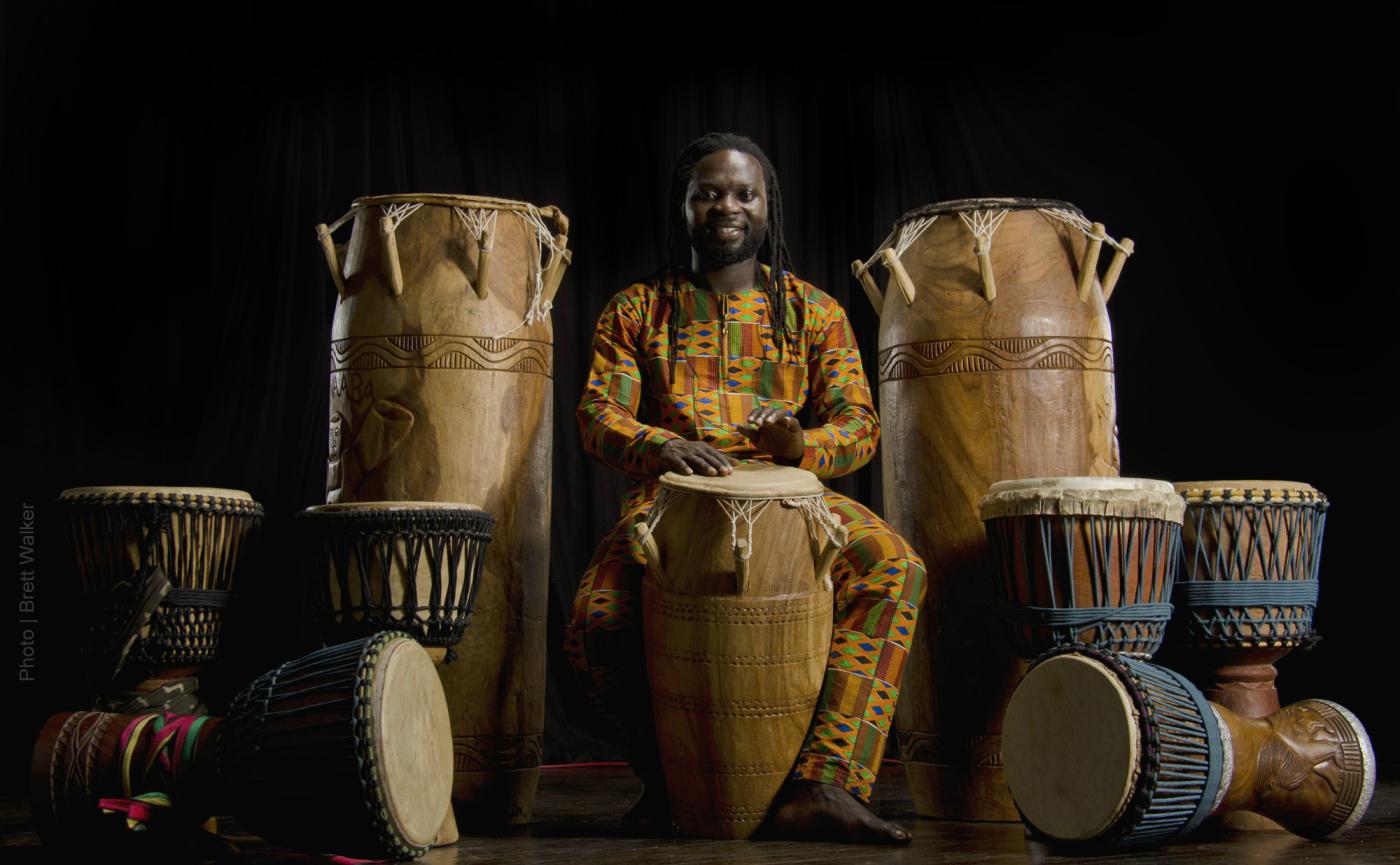 a sitting man dressed in West African garments is surrounded by a dozen drums of various sizes