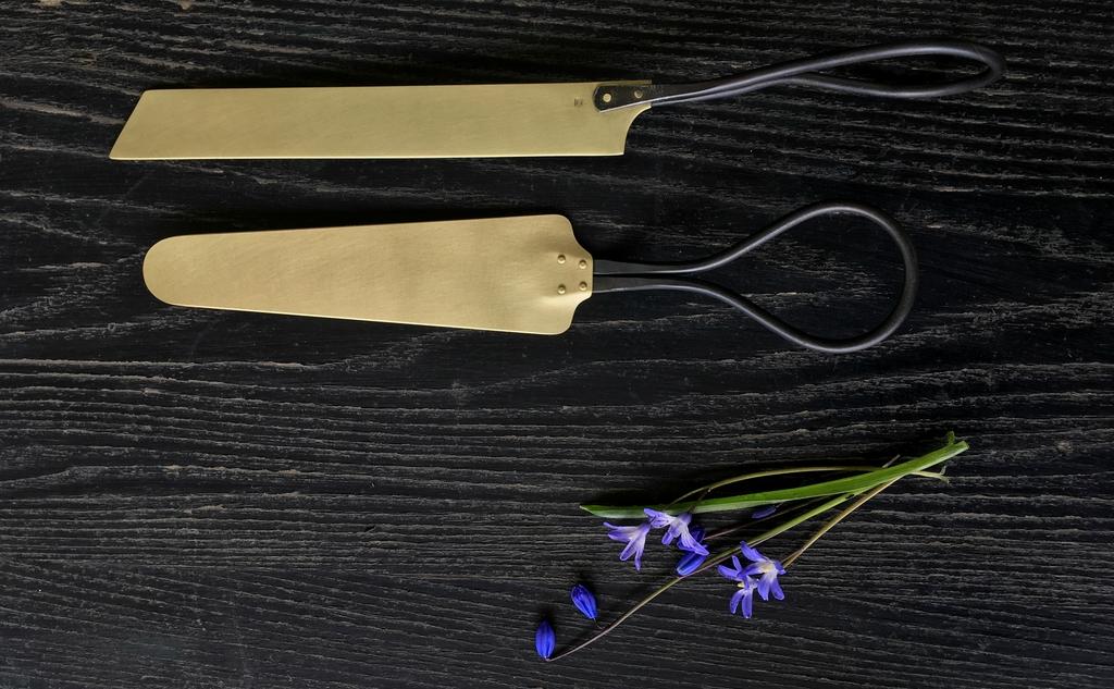 two rustic, golden metal cake knives are displayed on a dark wood table with a purple flower nearby