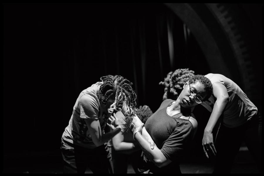 Black and white photo: three Black women lean on each other in a spotlight.