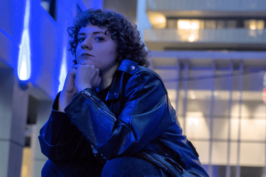 A person with curly hair in a leather jacket squats in the middle of the street.