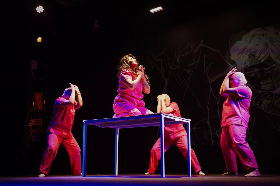 Three marked performers in prison jumpsuits surround a person in a jumpsuit who prays on top of a table.