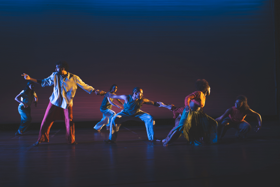 Indoors, on a stage, four performers hold hands forming a chain. There are two people behind them mid movement, and a woman in front.