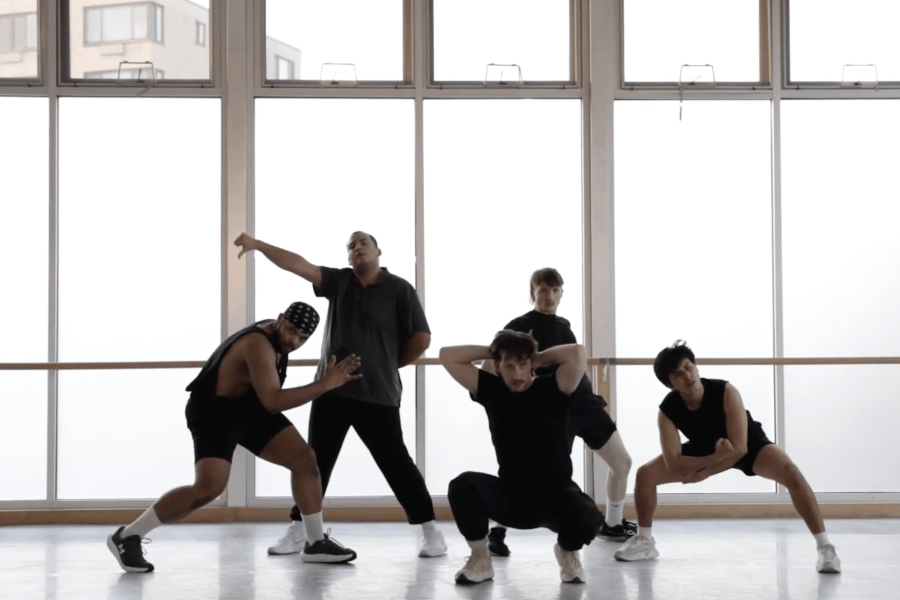 A group of five dancers wearing black poses in front of a studio wall.