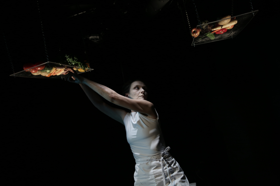 A white woman, in a white dress, pulls vegetables from a crate, hanging in the air. A second crate hangs on her other side.