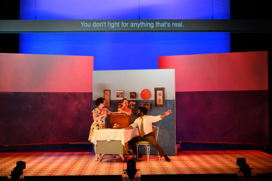 Three performers on a colorful set, with supertitles above reading “You don’t fight for anything that’s real.” One performer is holding a microphone up to another performer.