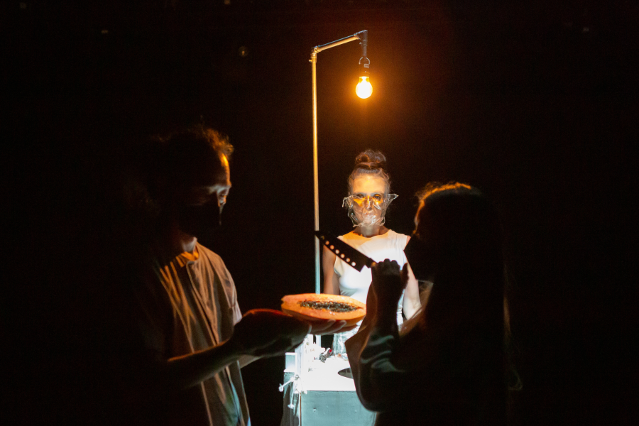 Three folks, under a floor lamp, wear masks and cut food with a big knife.