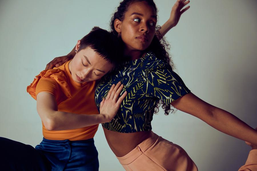 Two women perform in front of a white backdrop. One pushes into the other.