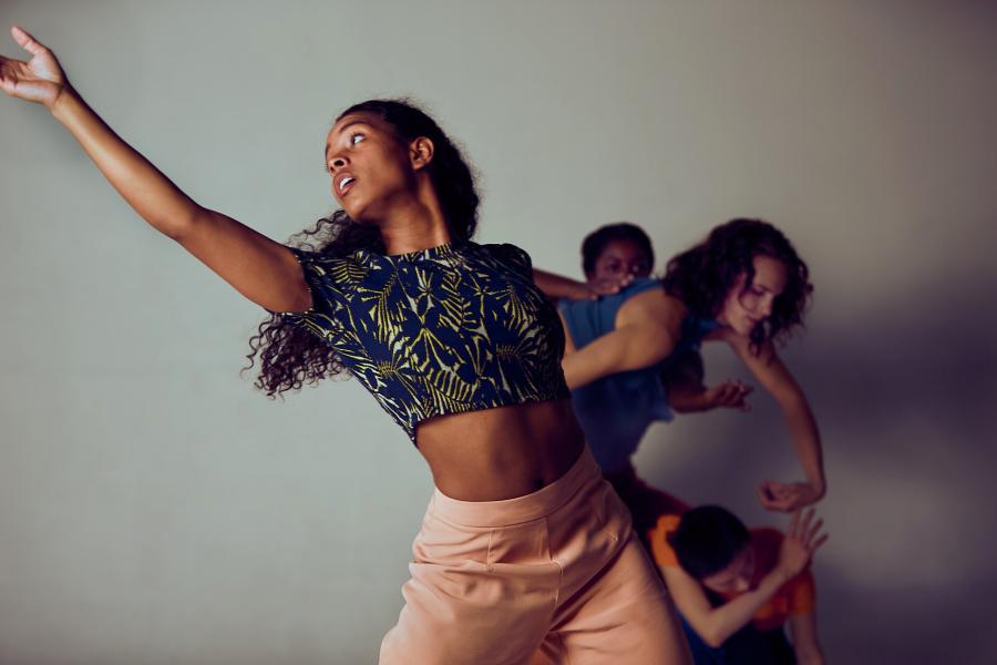 Four women perform in front of a white wall; they lean into each other and one reaches out in front of her.