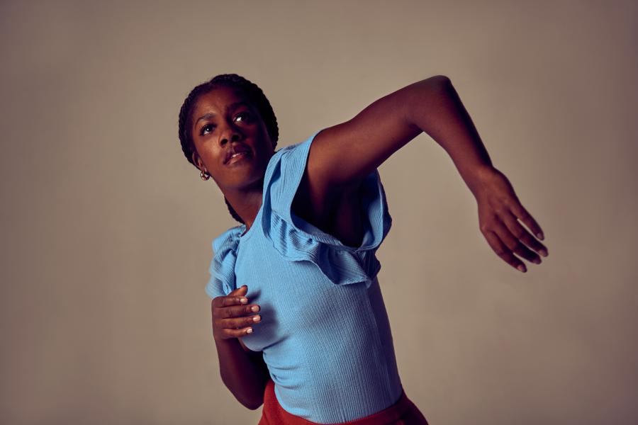 A Black woman holds her elbow up towards the ceiling. She wears a blue blouse with floppy sleeves.