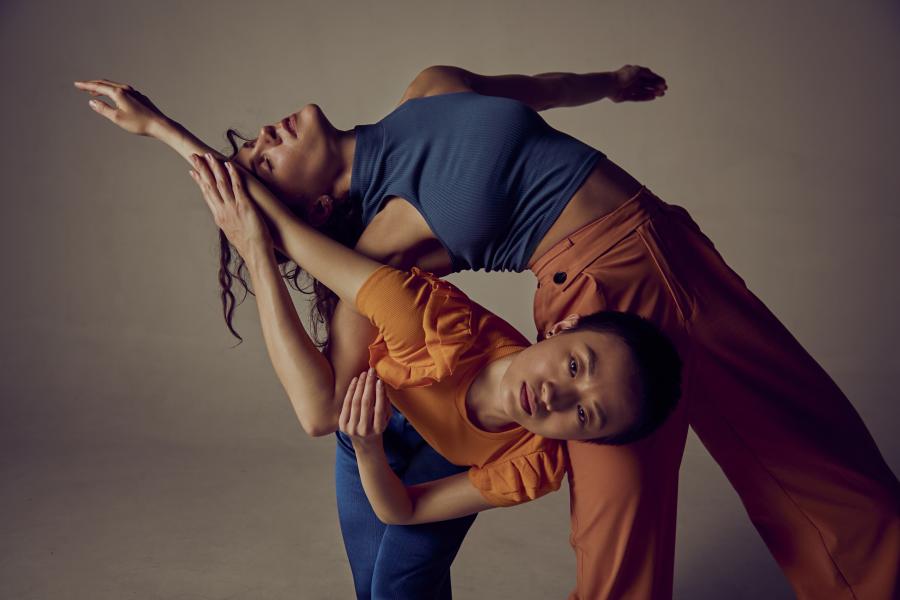 Two dancers, in front of a white backdrop, lean over and under each other. 