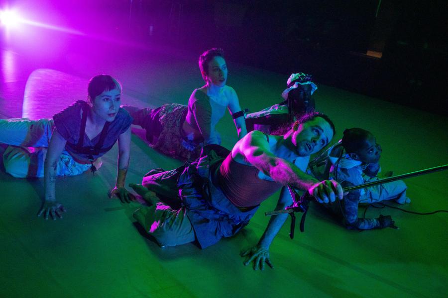 Laying on a stage, in green, purple, and blue light, five dancers crawl. One holds a pole that goes out of the frame.