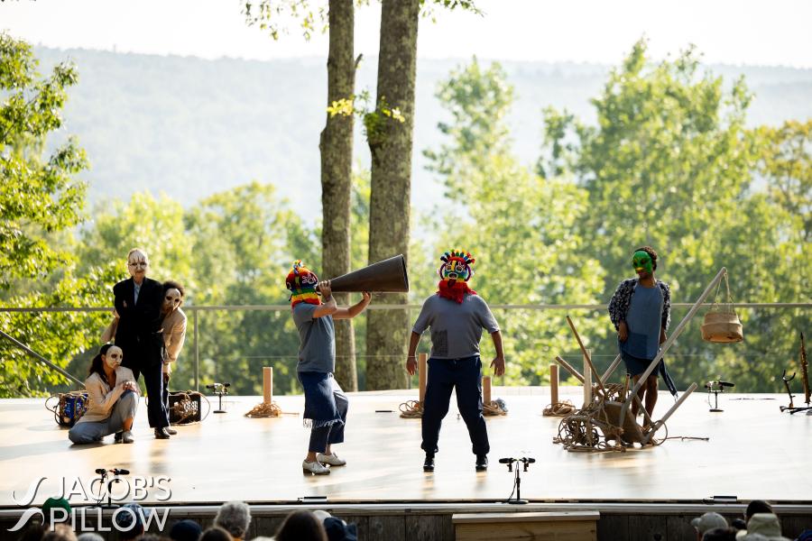 On a stage a group of masked performers observe a person in a bright mask.