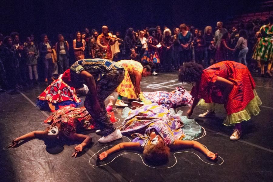 An audience watches in a Black box theater as four dancers draw body outlines around other dancers.