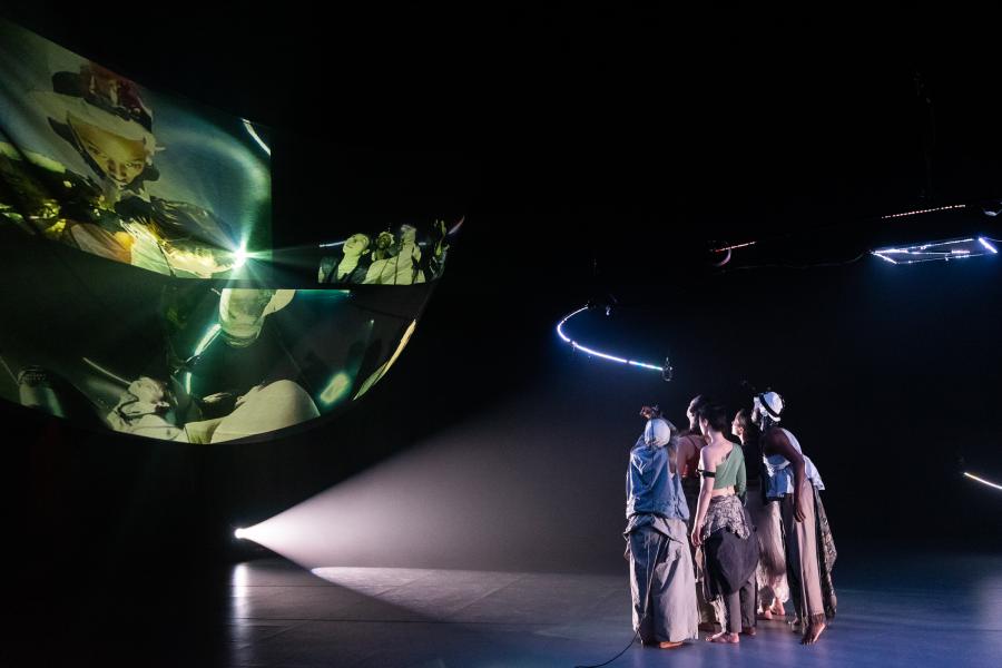 On a stage, a group of people look to a projection of a feminine person.