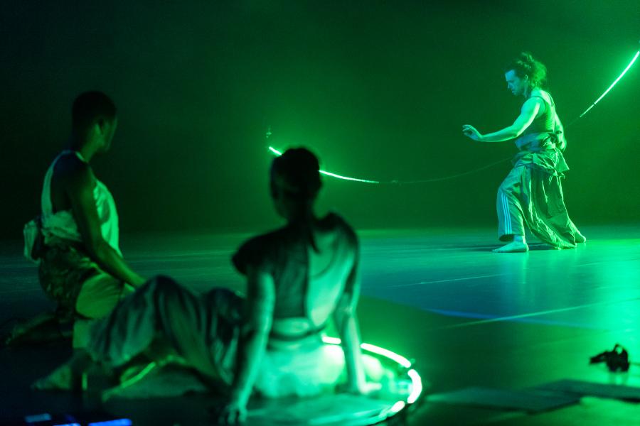 Two folks, sitting or kneeling on a stage, watch another person, on the stage and in green light, dance.