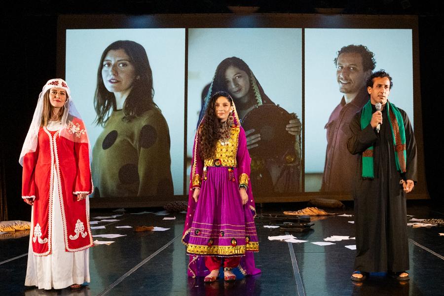 In front of screens with projections of each of the actor's headshots, three performers stand. Two women and a man, who holds a microphone.