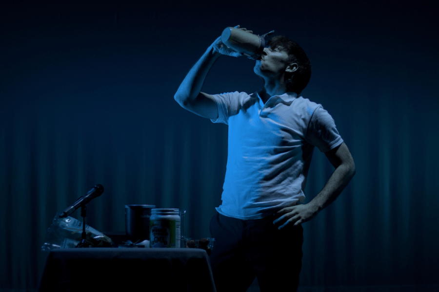 A person in a white polo shirt drinks from a mug in front of a dimly lit curtain.
