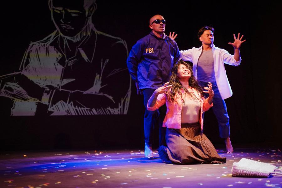 A person dressed like an FBI agent stands stoically behind two posing performers.