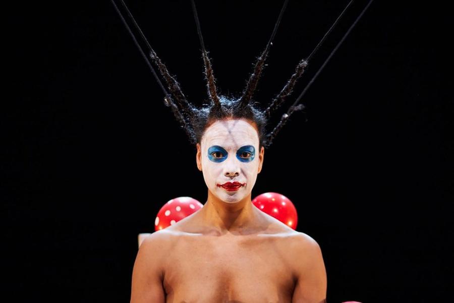 A woman, with her hair braided straight up, wears white face paint, with clownish blue eyes and red lips.