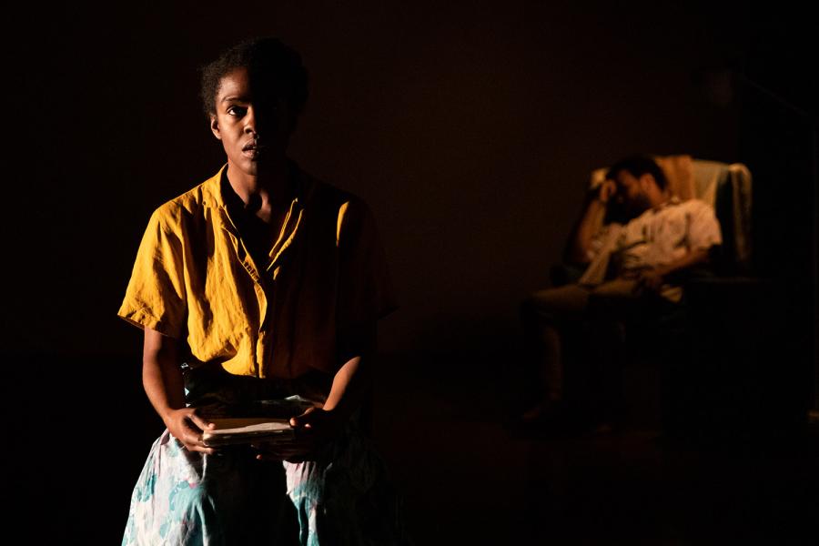 In the foreground, a Black woman in a spotlight, addresses the camera with papers in her hand. Behind her, a man sits in a chair, he leans and appears asleep.