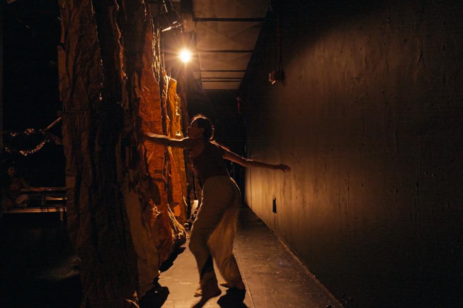 In a hallway, a dancer holds her hands against the walls. A spotlight catches her tank top and baggy pants.