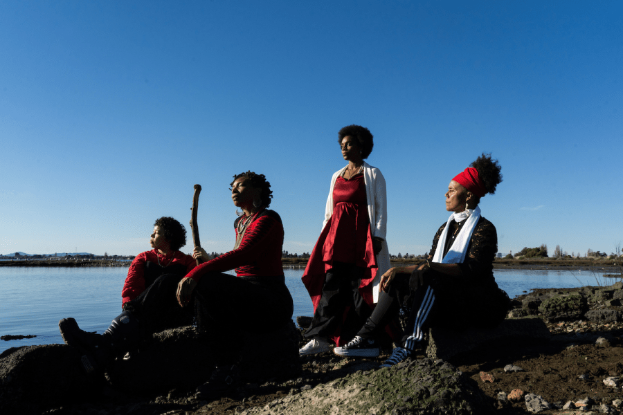 Four people are outside by a waterfront, three of them sit and one of them stands.