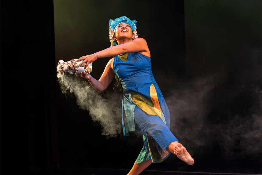 On a stage, a woman wears blue and wears a headdress while holding something that smokes like sage.