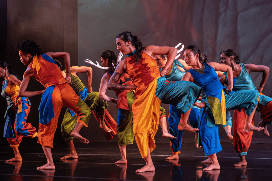 On a stage and in formation, folks in oranges and blues lift one leg and lean to the other side.