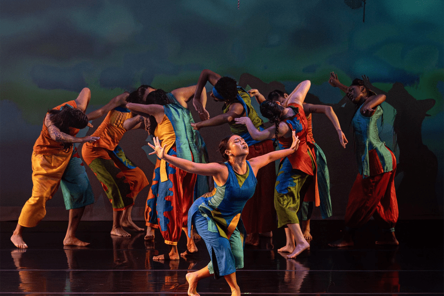 On stage, one woman in blue extends her arms almost to bow while behind her the company leans in together as if in a huddle.
