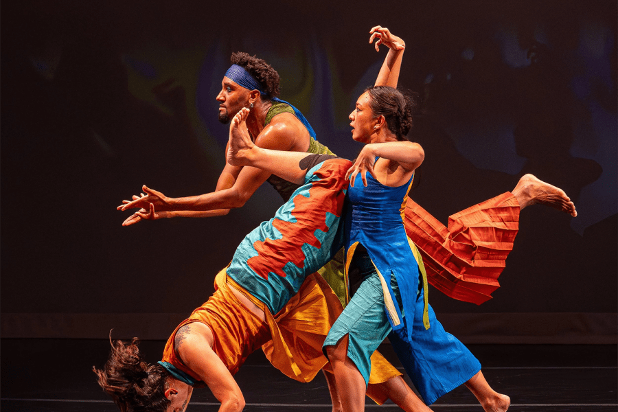 On a stage, two folks in blue help a person in orange doa  cartwheel.