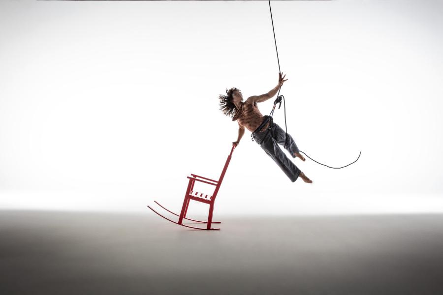 A person suspended in midair by a harness reaches upward with their left hand while touching a red rocking chair with their right.