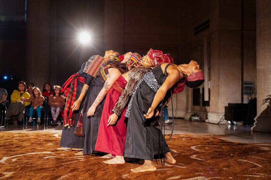 On a stage, folks wear denim, black, and red. They lean back in a row and in spotlight.
