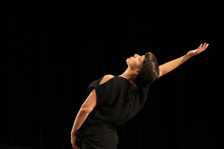Against a jet black background, a Native woman with medium brown skin, dark brown straight hair in a single loose braid, and regal facial expression arches back in a dancerly position with her right arm raised high, looking up. She wears a black outfit with a sleeveless top